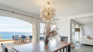 Un salon moderne doté d’une grande fenêtre donnant sur l’océan, d’un coin repas avec une table en marbre et d’un lustre élégant.