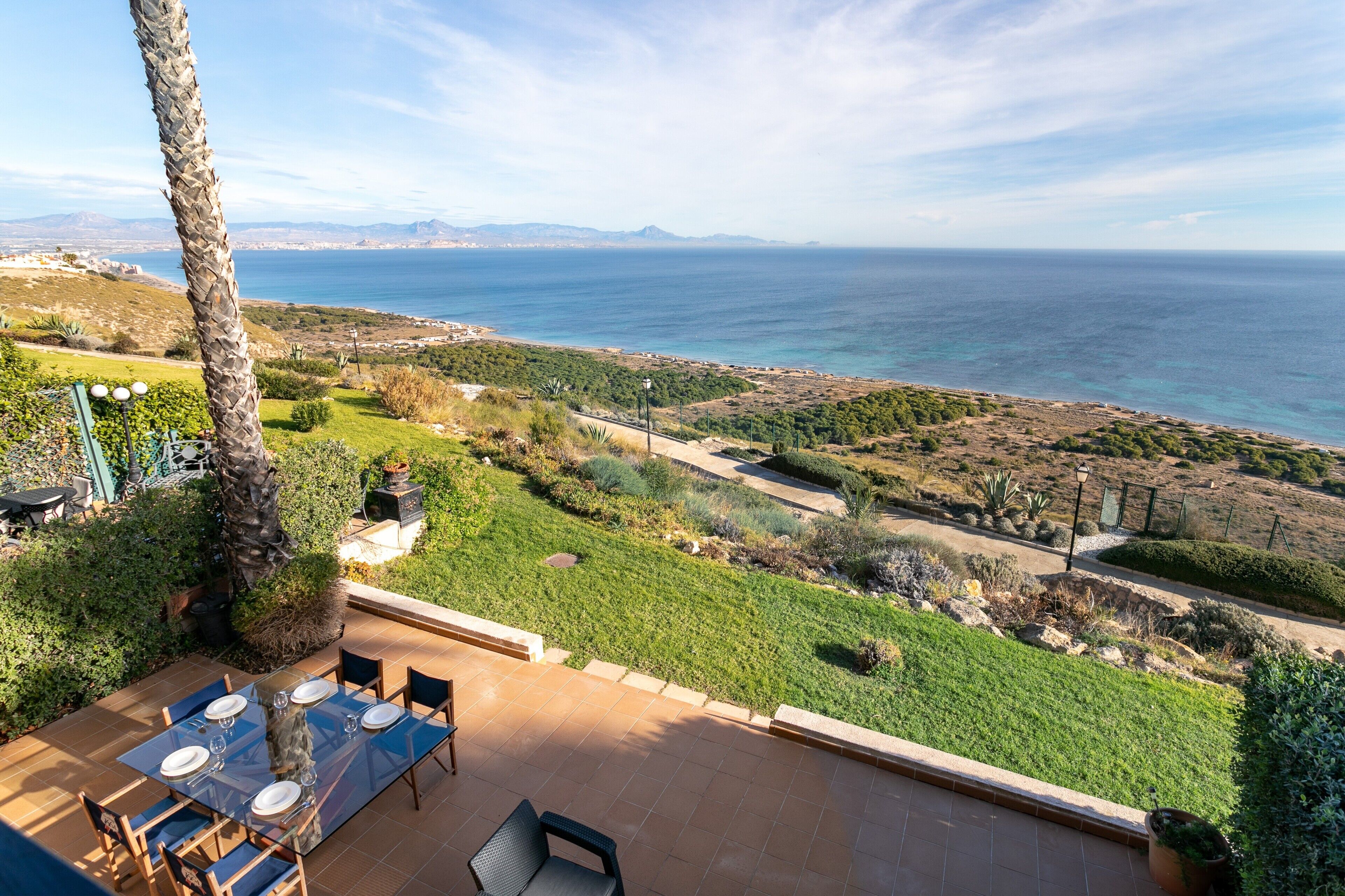 Une terrasse avec une table dressée pour le repas, offrant une vue sur un paysage côtier avec la mer et des montagnes lointaines à l’horizon.