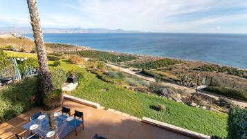 Une terrasse avec une table dressée pour le repas, offrant une vue sur un paysage côtier avec la mer et des montagnes lointaines à l’horizon.