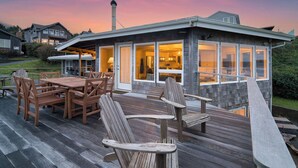 A wooden deck with outdoor furniture, a covered patio area, and a view of the house's interior through large windows. - Sanctuary by the Sea by Avantstay Waterfront + Views (Arch Cape)