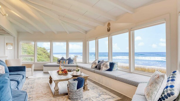 A sunroom with a view of the ocean, featuring a blue sofa, a wooden coffee table, and a bench with cushions. - Sanctuary by the Sea by Avantstay Waterfront + Views (Arch Cape)