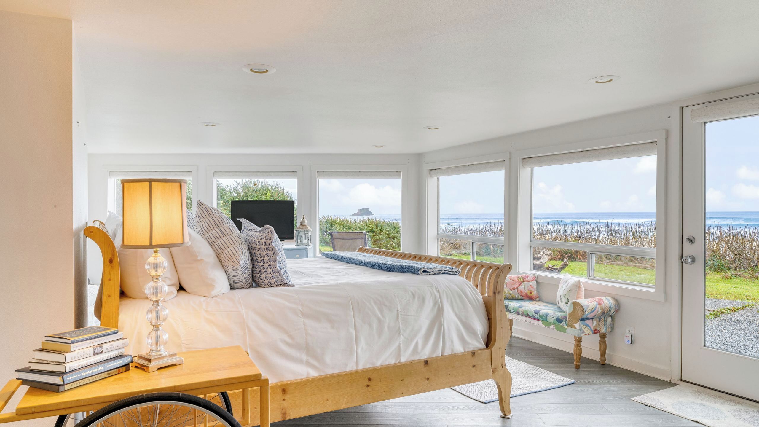 Un dormitorio con cama, mesita de noche con libros, lámpara y vistas al océano.