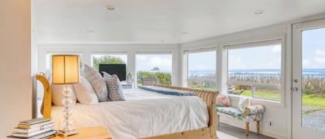 Un dormitorio con cama, mesita de noche con libros, lámpara y vistas al océano.