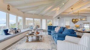 Ein geräumiges Wohnzimmer mit großem Fenster zum Strandblick, ein blaues Sofa, ein Couchtisch und ein Essbereich mit klarem Glasgeschirr.