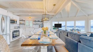 A modern kitchen with white cabinetry, a dining area with a wooden table set for six, and a large window overlooking the ocean. - Sanctuary by the Sea by Avantstay Waterfront + Views (Arch Cape)
