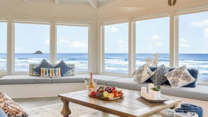 A room with a large window overlooking the ocean, a wooden coffee table with a fruit and cheese board, and a seating area with patterned cushions. - Sanctuary by the Sea by Avantstay Waterfront + Views (Arch Cape)