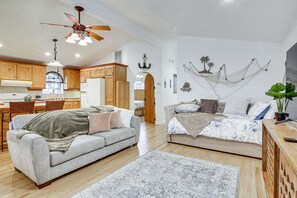 A spacious living area with a grey sofa, a beige sofa, and a wooden coffee table. There is a ceiling fan, a chandelier, and a kitchen area with wooden cabinets and a window.