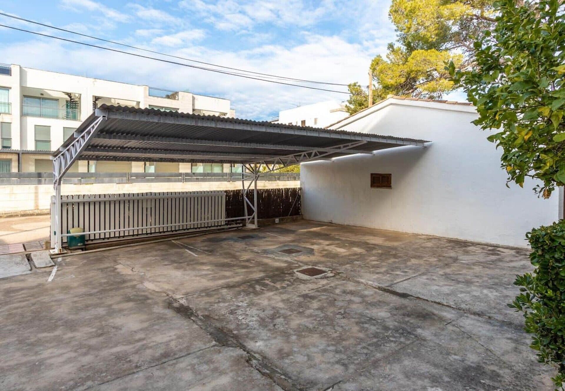 A covered outdoor area with a concrete floor, a metal fence, and a white building in the background.
