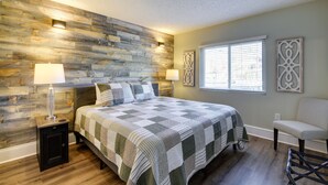 Une chambre avec un mur en bois, un lit recouvert d’une courtepointe, deux lampes de chevet, une chaise et une fenêtre avec des stores.