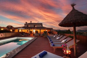 A poolside area with lounge chairs, a thatched umbrella, and a house in the background. - Country House 'Ca Sou' with Private Pool, Wi-Fi and Air Conditioning (Alcudia)