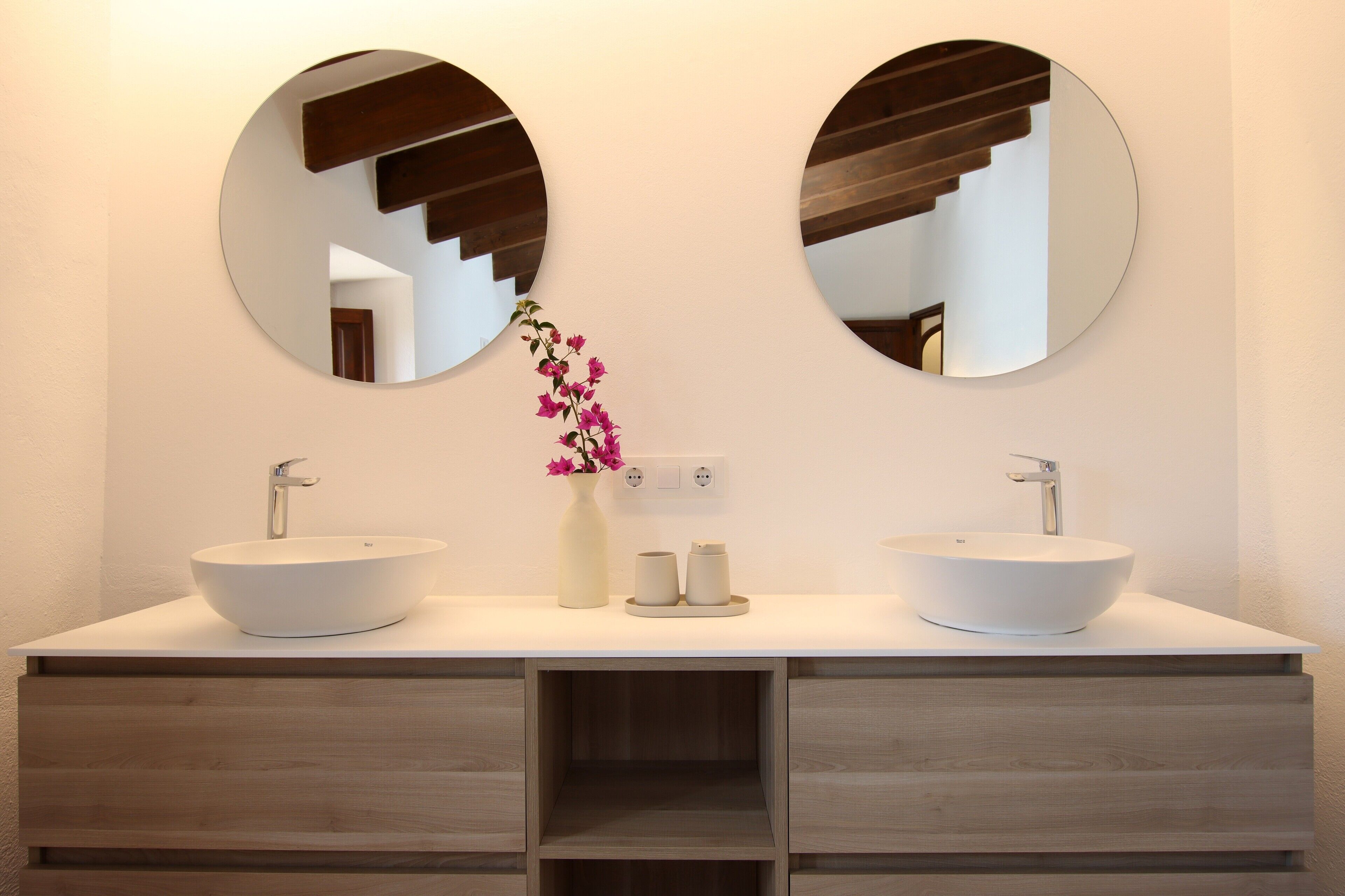 Une salle de bain avec deux lavabos, un vase rempli de fleurs et deux miroirs ronds placés sur un comptoir blanc.