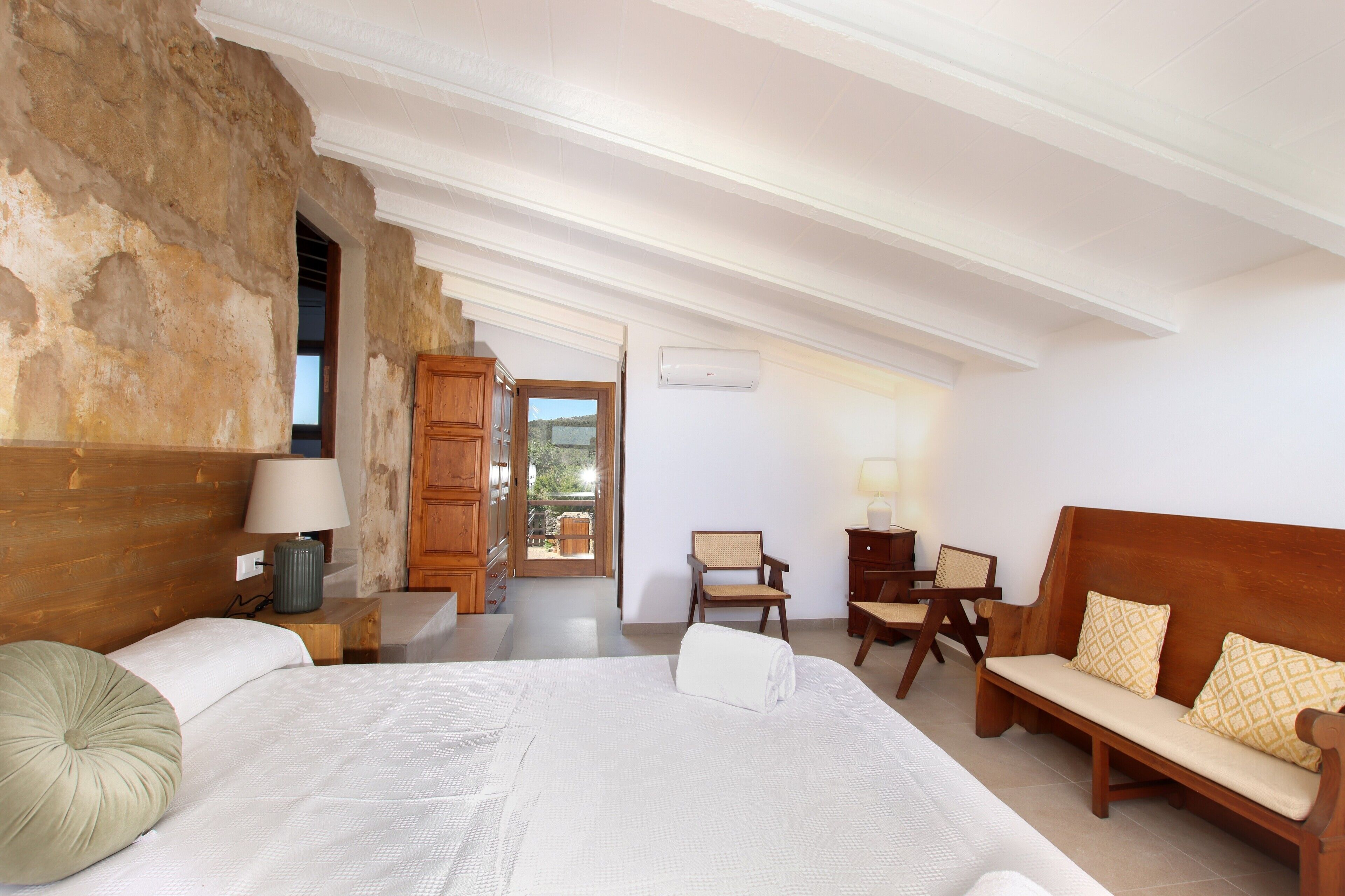 A bedroom with a large bed, wooden headboard, a wooden bench, a chair, and a view of the outdoors.