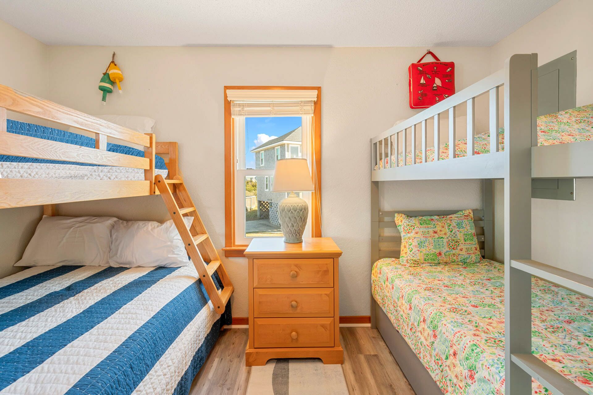 A bedroom with bunk beds, a wooden dresser, a window with a view of houses, and a red wall decoration.