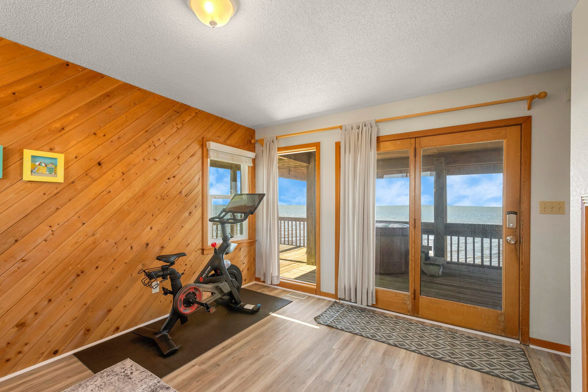 Una stanza con pavimento in legno, una cyclette, vista sull'oceano tramite una porta a vetri scorrevoli e un piccolo quadro appeso al muro.