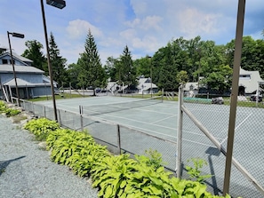 A tennis court with a chain-link fence, surrounded by greenery and residential buildings. - BearHug AC, Pool, Hot Tub, Tennis, Close to Resort (Killington)