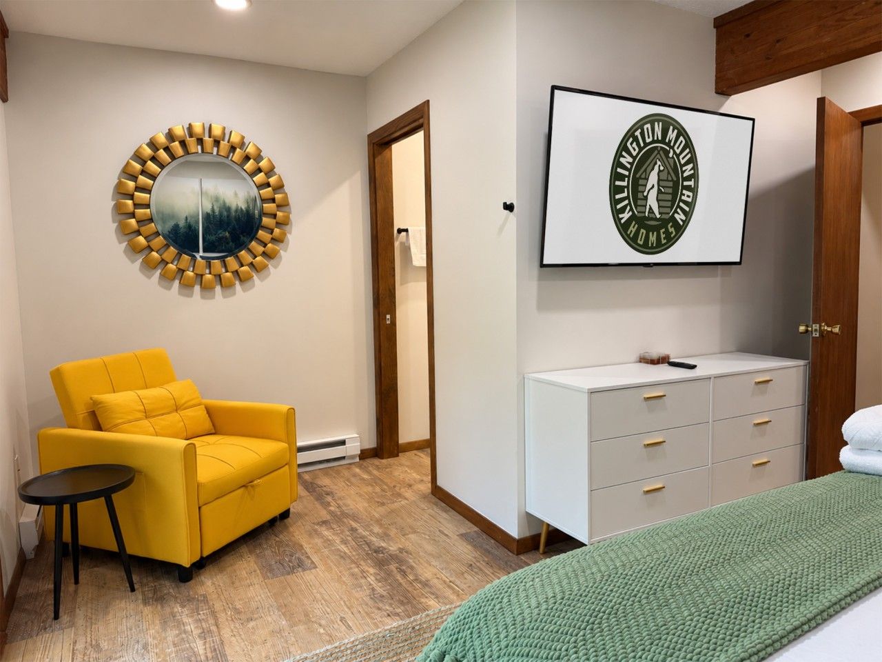 A room with a yellow armchair, a small round table, a white dresser, and a framed picture on the wall.