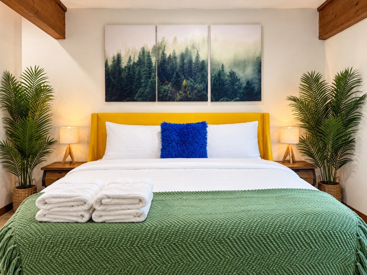 A hotel room with a bed, green textured bedspread, white pillows, a blue accent pillow, and a forest-themed wall art.