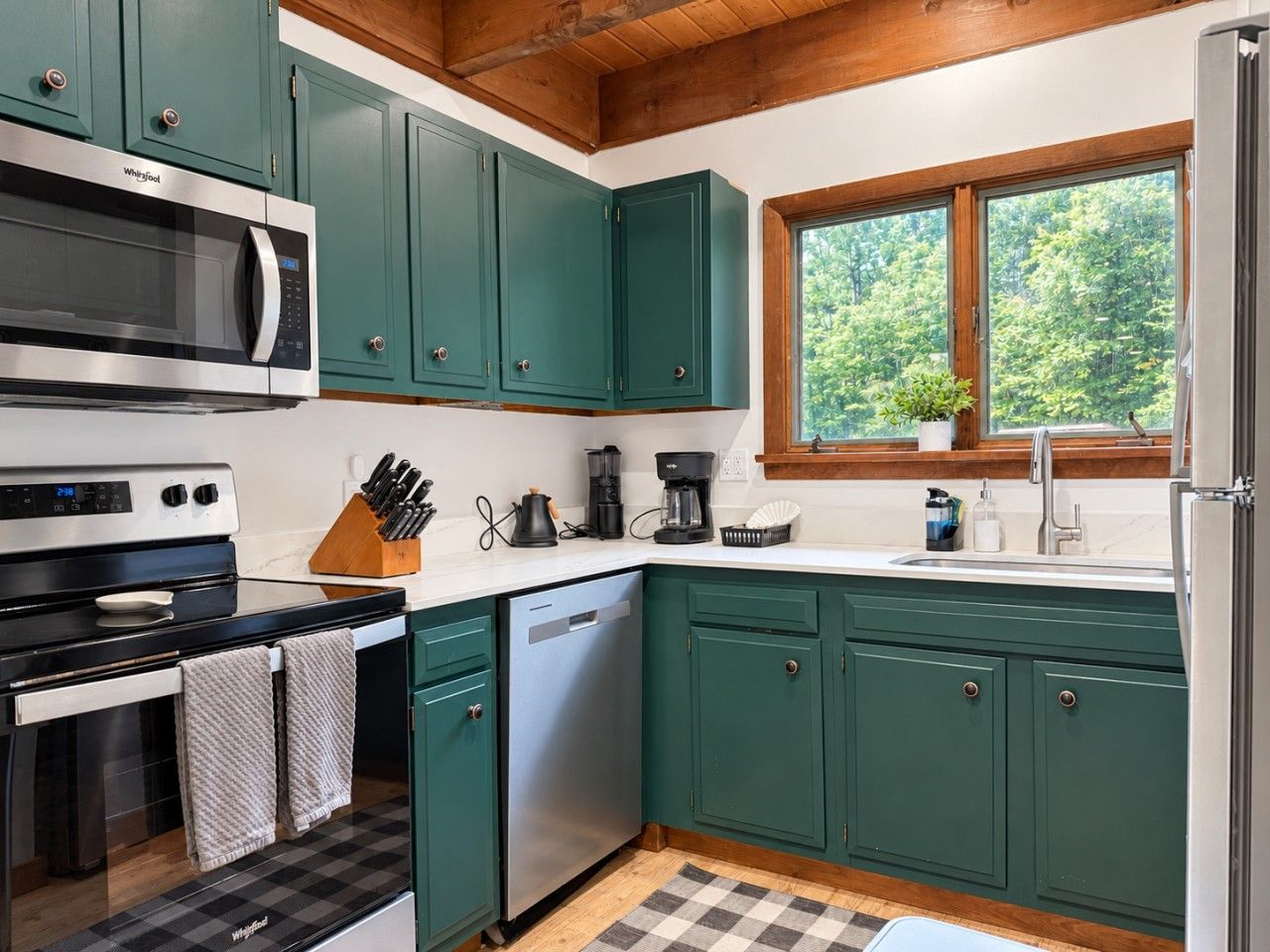 A kitchen with green cabinets, a microwave, stove, dishwasher, and a window with a view of trees.