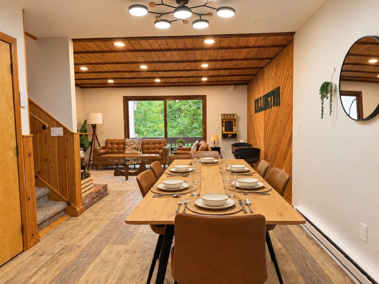 A dining area with a wooden table set for four, a staircase, and a view of greenery outside.