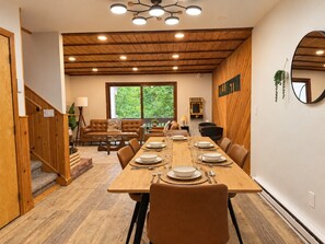 Un comedor con una mesa de madera dispuesta para cuatro, una escalera y vistas al exterior con vegetación.