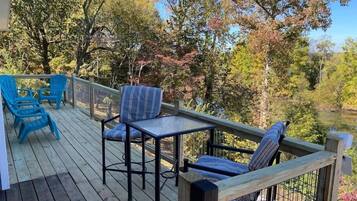 A wooden deck with outdoor seating, including chairs and a table, overlooking a body of water and surrounded by trees.