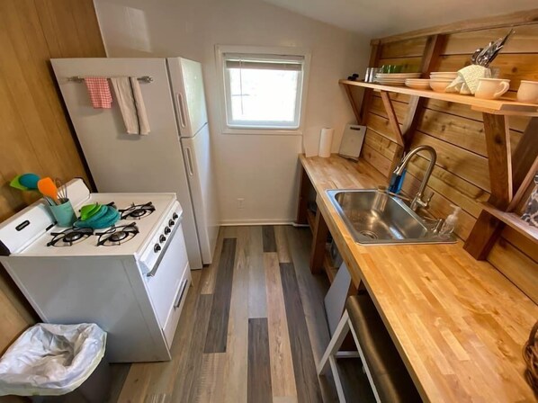 A compact kitchen with a gas stove, sink, wooden shelves, and a window.
