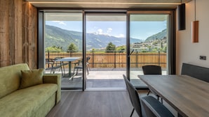 A modern room with a wooden wall, a sofa, a table, and a view of mountains and a town through large glass doors. - Apartment 'Köfele Lodge Sepp' with Mountain View, Shared Pool and Wi-Fi (Partschins)