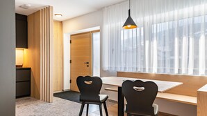 Une pièce moderne avec un bureau en bois, deux chaises noires, un luminaire suspendu et une grande fenêtre ornée de rideaux légers.