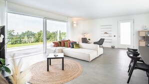 Ein modernes Wohnzimmer mit einem weißen Sofa, einem runden Teppich, einem Esstisch mit Stühlen und Blick auf Grün durch große Fenster.