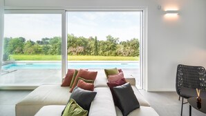 Ein modernes Wohnzimmer mit einer großen Glasschiebetür, die zu einem Pool führt, einer Couch mit Kissen und einem schwarzen Rattan-Sessel.