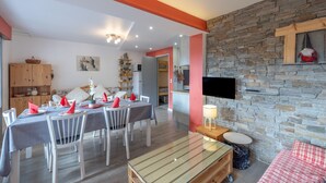 A dining area with a table set for four, a sofa, a television mounted on a stone wall, and a wooden cabinet. - Apartment 'Appartement Spacieux 6 Personnes' with Mountain View, Private Terrace and Wi-Fi (LE GRAND BORNAND)