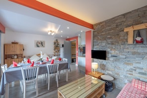 A dining area with a table set for four, a sofa, a television mounted on a stone wall, and a wooden cabinet. - Apartment 'Appartement Spacieux 6 Personnes' with Mountain View, Private Terrace and Wi-Fi (LE GRAND BORNAND)
