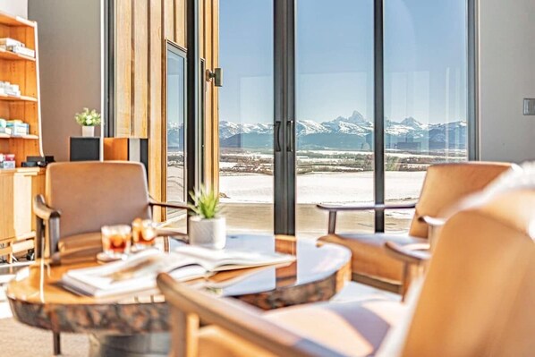 A modern hotel room with a large window offering a mountain view. There are two leather armchairs and a small table with a book and a glass of water.