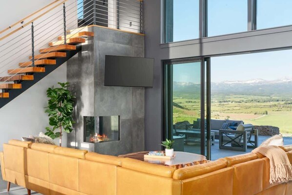 A modern living room with a sofa, a fireplace, and a large window overlooking a scenic landscape. - Basecamp Stays: Lux Retreat, Epic Views, & Hot Tub (Tetonia)