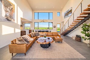 A spacious living room with a large sectional sofa, a round coffee table, and a view of the outdoors through large windows.