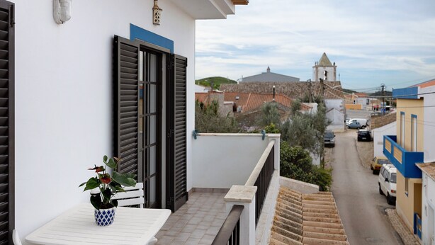 A balcony with a potted plant, a white table, and a view of a street with buildings and a church tower. - Holiday Home 'Brita' with Private Terrace, Balcony and Wi-Fi (Raposeira)