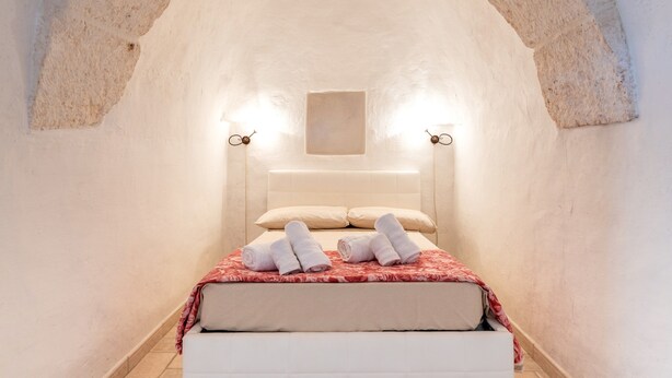 Une pièce confortable à l’atmosphère chaleureuse, avec un plafond en arc, un lit, deux lampes fixées au mur et une petite fenêtre carrée.