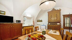 A dining area with a wooden table, chairs, a fruit basket, a television, and a kitchen in the background.