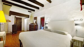 A bedroom with a large bed, wooden beams on the ceiling, a dresser, and a lamp.