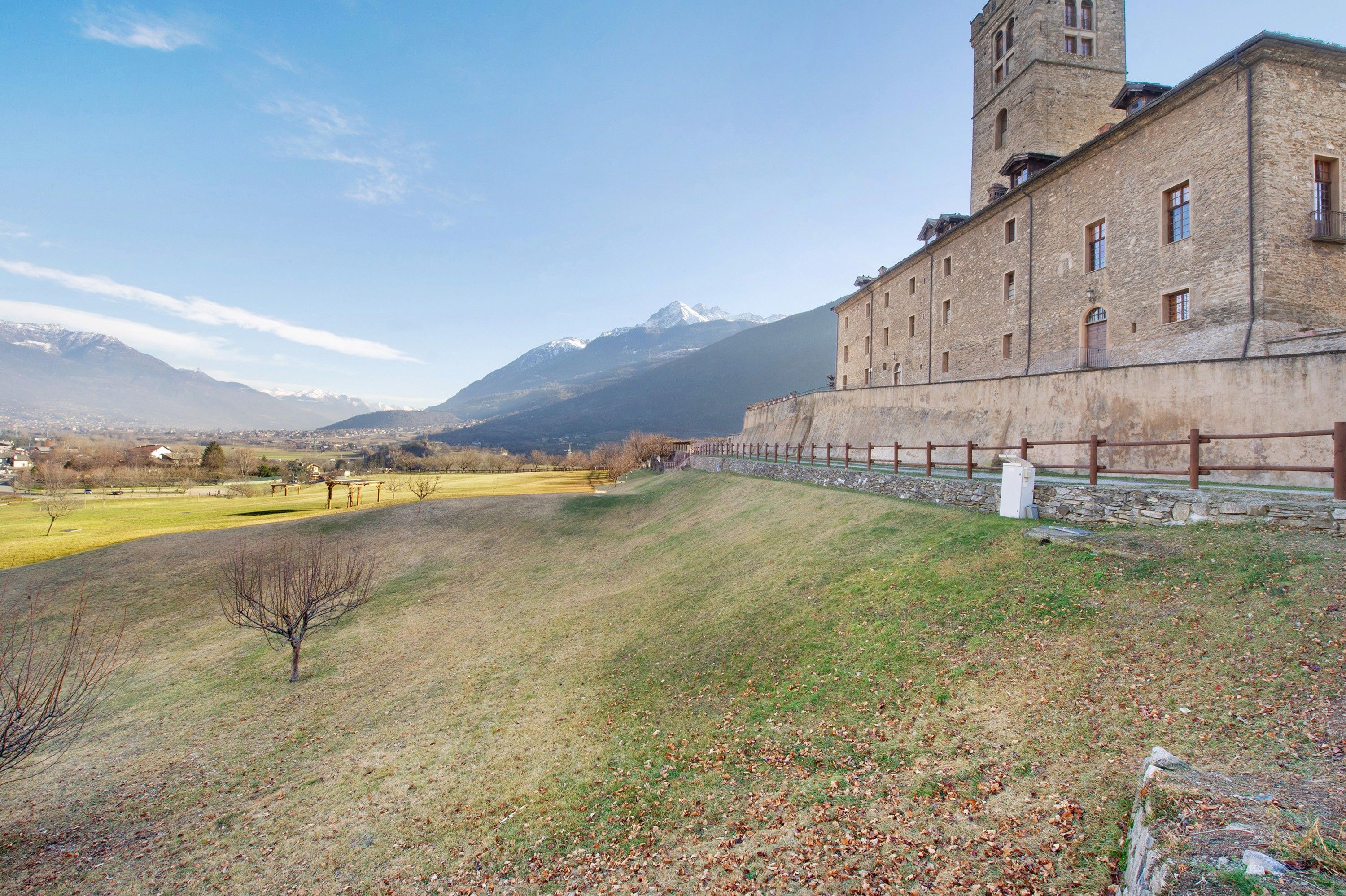 Un edificio histórico con una torre, una colina cubierta de pasto y montañas al fondo.