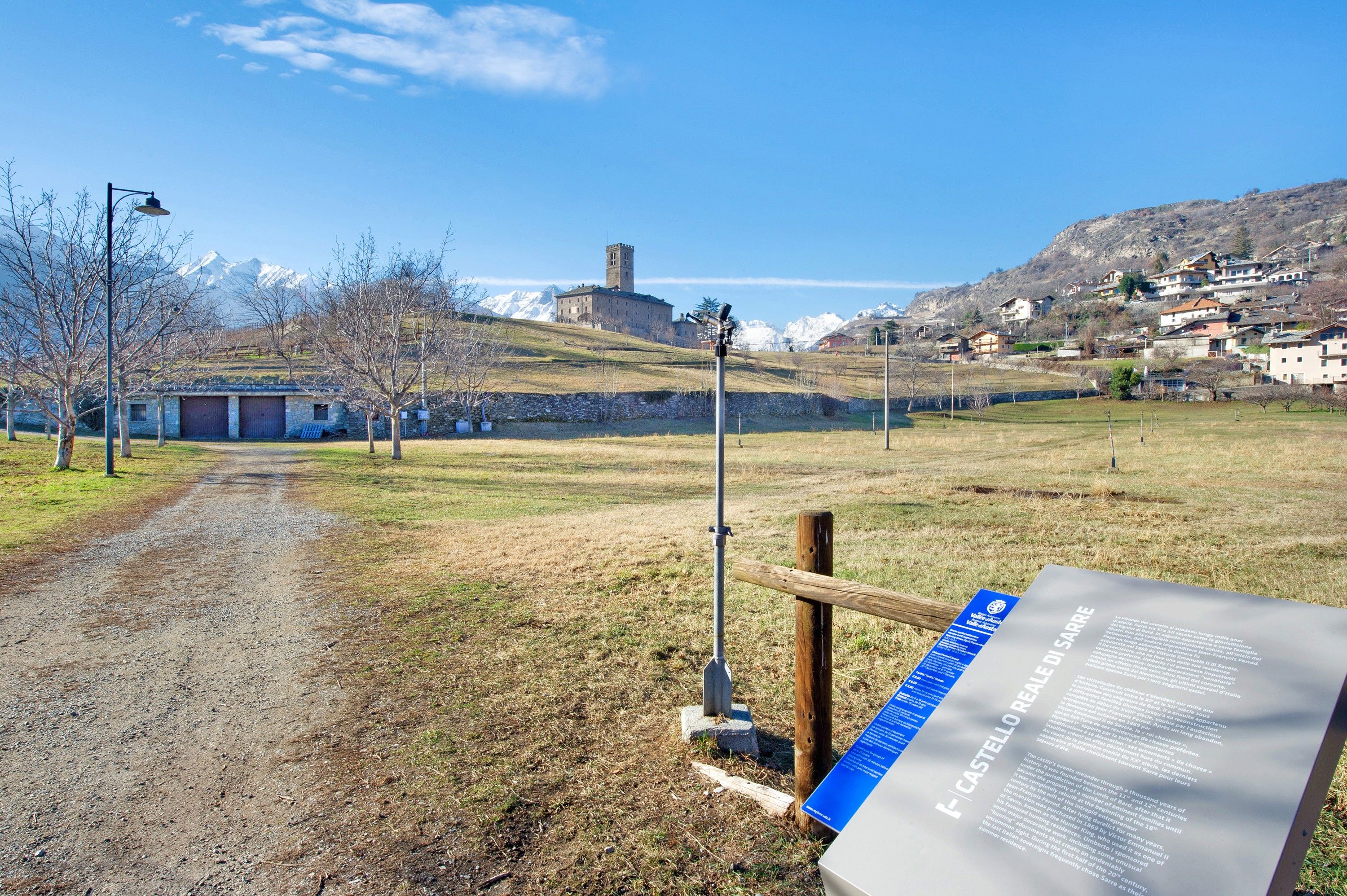 Un paysage rural avec un chemin de terre menant à un bâtiment historique, un panneau indicateur et une chaîne de montagnes en arrière-plan.
