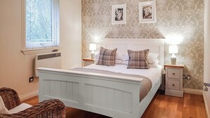 A bedroom with a bed, two bedside tables, a window with blinds, and a wall with floral wallpaper. - Leven Lodge 3 - UK46045 (Loch Lomond)