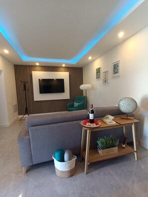 A modern living room with a grey sofa, a flat-screen TV mounted on the wall, a green armchair, a wooden coffee table with a bottle of wine, and a decorative lamp.