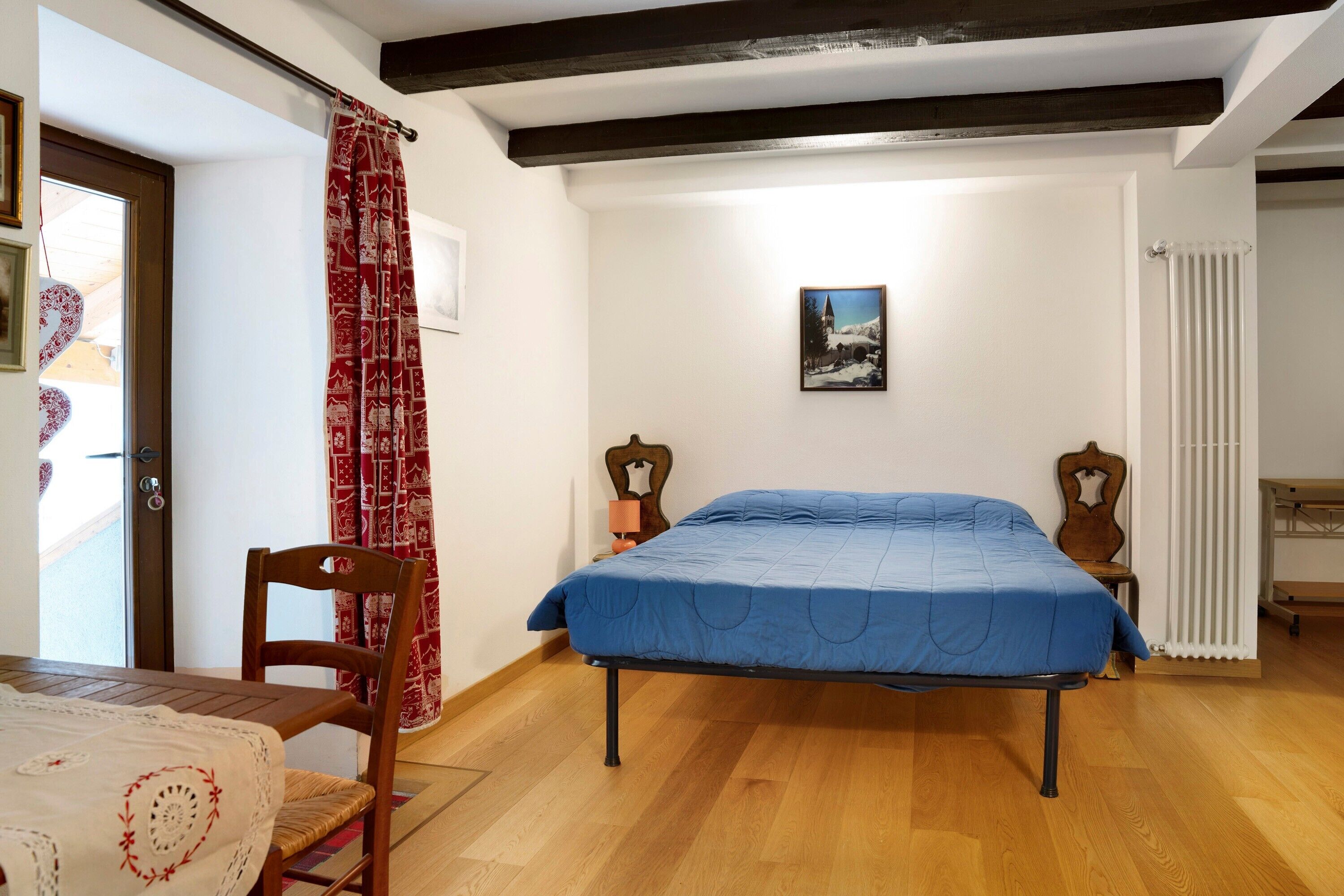 A bedroom with a bed, wooden floor, a chair, a table, a red patterned curtain, a picture on the wall, and a radiator.