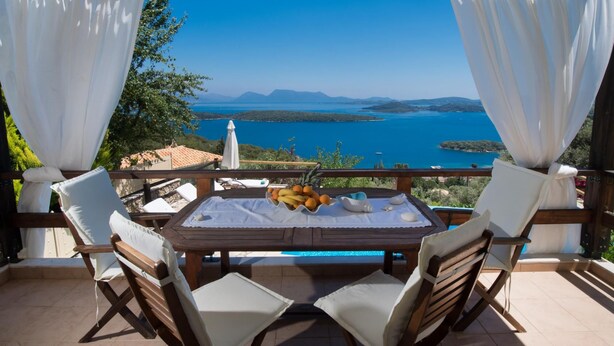 A wooden table with a white tablecloth, surrounded by four wooden chairs, overlooking a scenic view of a blue sea and distant mountains. - Stunning Seaviews Pool Beach - Pasithea (Lefkada)
