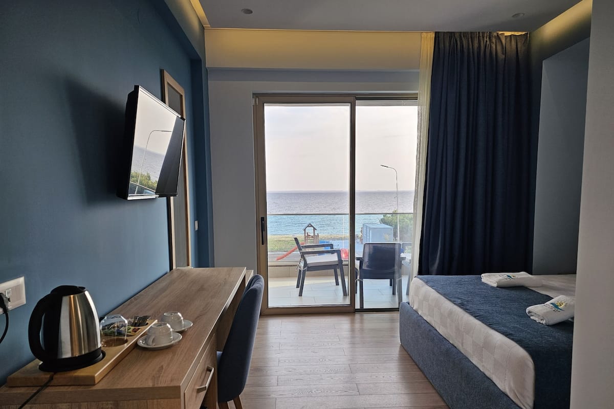 A hotel room with a bed, a desk with a kettle and cups, a TV, and a view of the sea through a glass door.