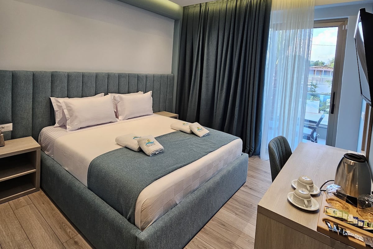 A modern hotel room with a large bed, a desk with a kettle, and a view of the outdoors.