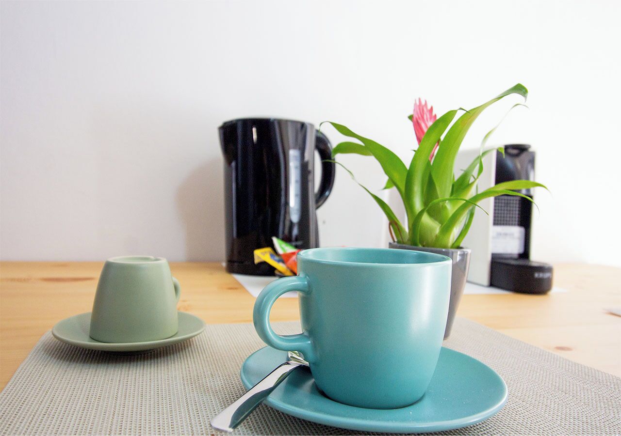A teal mug on a matching saucer with a spoon, a small green cup, and a potted plant with pink flowers on a wooden table.