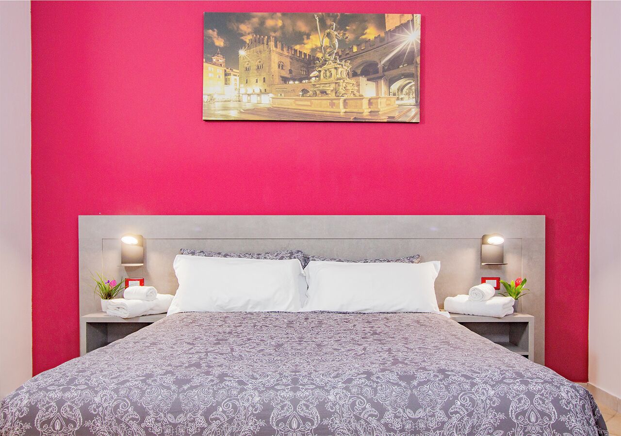 Una habitación de hotel con una cama grande, dos mesitas de noche, una lámpara de noche, un cuadro sobre la cama y una pared roja.