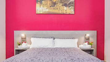 Una habitación de hotel con una cama grande, dos mesitas de noche, una lámpara de noche, un cuadro sobre la cama y una pared roja.